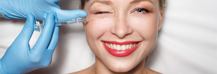 Blonde lächelnde Frau liegt auf einem Behandlungsstuhl und zwinkert während ihre Augenfalten unterspritzt werden