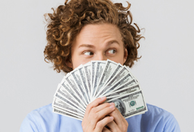 Junger Mann mit lockigem Haar hält Fächer aus Geldscheinen vor sein Gesicht