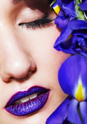 Nahansicht einer Frau mit geschlossenen Augen und lila farebenen Lippen. Blüten bedecken eine Gesichthälfte.