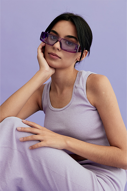 stylische Frau mit violetter Sonnenbrille mit lila Tank Top und Rock sitzen auf pastellviolettem Hintergrund, zeigt entspannte Pose und moderne Mode